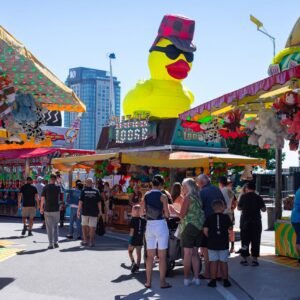 Colorful summer carnival scene with a giant rubber duck and game stalls under clear blue sky.